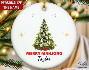 May include: White ceramic Christmas ornament with a gold string for hanging. The ornament features a green Christmas tree decorated with Mahjong tiles. The words "MERRY MAHJONG" are in red, with a personalized name below. The text "PERSONALIZE THE NAME" is at the top.