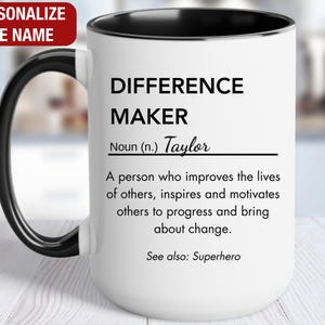 May include: White ceramic mug with a black rim and handle. The mug has a black inscription that reads "Difference Maker" and defines the term as "A person who improves the lives of others, inspires and motivates others to progress and bring about change." The inscription also includes the name "Taylor" and the phrase "See also: Superhero".