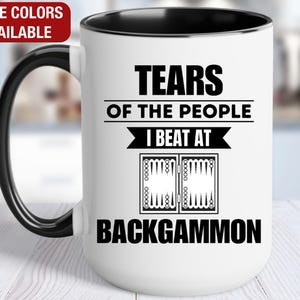 May include: White ceramic coffee mug with a black rim and handle. The mug features a black and white graphic of a backgammon board and the text "Tears of the people I beat at Backgammon".
