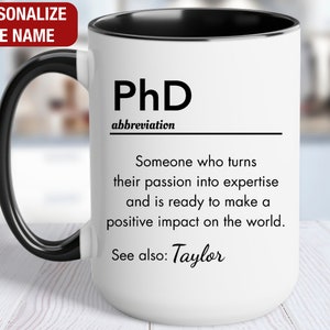 May include: White ceramic coffee mug with a black handle and black rim. The mug has a black and white graphic that reads "PhD abbreviation Someone who turns their passion into expertise and is ready to make a positive impact on the world. See also: Taylor"