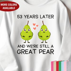 May include: White sweatshirt with the text "53 YEARS LATER AND WE'RE STILL A GREAT PEAR". The design features two green pear illustrations with smiling faces and small red hearts. A red oval label says "MORE COLORS AVAILABLE".