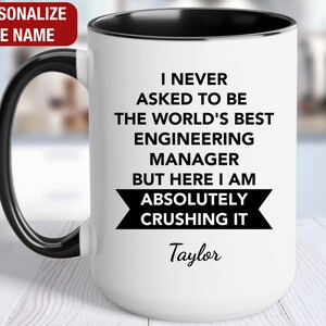May include: White ceramic coffee mug with a black rim and handle. The mug has black text that reads "I never asked to be the world's best engineering manager but here I am absolutely crushing it." The text "Taylor" is printed below the text.