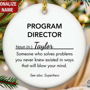 May include: A white ceramic Christmas ornament with the text "PROGRAM DIRECTOR" and the name "Taylor". The ornament includes a definition and the words "See also: Superhero". A gold string is attached for hanging.