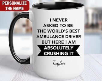 May include: White ceramic coffee mug with a black rim and handle. The mug has black text that reads "I never asked to be the world's best ambulance driver but here I am absolutely crushing it." The name "Taylor" is printed below the text.