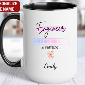 May include: White ceramic mug with a black rim and handle. The mug has a pink and blue progress bar with the text "Engineer in progress..." and a floral design. The name "Emily" is printed below the progress bar.