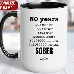 May include: White ceramic mug with a black rim and handle. The mug has black text that reads "30 years", "360 months", "1560 weeks", "10950 days", "262800 hours", "15768000 minutes", "946080000 seconds", "SOBER", and "Taylor".
