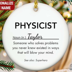 May include: A white ceramic Christmas ornament with the word "PHYSICIST" in black. Below, the name "Taylor" is written, followed by a definition. The ornament has a gold string for hanging and the text "PERSONALIZE THE NAME" in a red box.