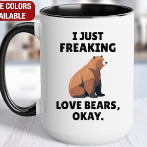 May include: White ceramic mug with a black handle and rim. The mug features the text "I JUST FREAKING LOVE BEARS, OKAY." and a brown bear illustration. The words "MORE COLORS AVAILABLE" are in a red box.