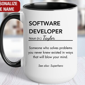 May include: White ceramic mug with a black handle and rim. The mug has a black and white design that reads "SOFTWARE DEVELOPER Noun (n.) Taylor Someone who solves problems you never knew existed in ways that will blow your mind. See also: Superhero"