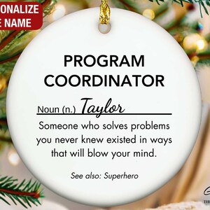 May include: A white ceramic Christmas ornament with gold hanging string. The ornament reads "PROGRAM COORDINATOR" with the name "Taylor" below. The text also includes a definition and the words "See also: Superhero."