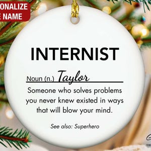 May include: A white ceramic Christmas ornament with the word "INTERNIST" in bold black letters. Below, the name "Taylor" is written in cursive. The ornament includes the text "Someone who solves problems you never knew existed..." and "See also: Superhero."