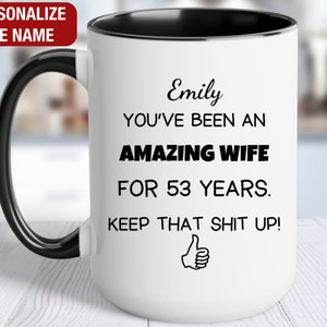May include: White ceramic coffee mug with a black handle and black rim. The mug has black text that reads "Emily You've Been An Amazing Wife For 53 Years. Keep That Shit Up!" with a thumbs up icon.