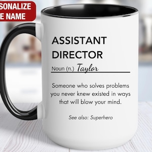 May include: White ceramic mug with a black rim and handle. The mug has a black and white design that reads "Assistant Director Noun (n.) Taylor Someone who solves problems you never knew existed in ways that will blow your mind. See also: Superhero"