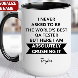 May include: White ceramic coffee mug with a black rim and handle. The mug has black text that reads "I never asked to be the world's best QA tester but here I am absolutely crushing it". The name "Taylor" is printed below the text.