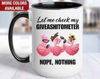 May include: White ceramic coffee mug with a black handle and rim. The mug has a black and white graphic of three pink flamingos with the text "Let me check my GIVEASHITOMETER NOPE, NOTHING".