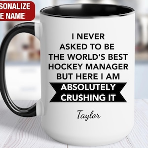 May include: White ceramic mug with a black handle and rim. The mug features the text "I NEVER ASKED TO BE THE WORLD'S BEST HOCKEY MANAGER BUT HERE I AM ABSOLUTELY CRUSHING IT" and the name "Taylor".