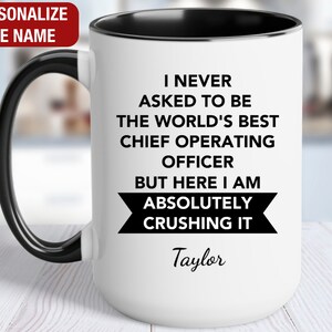 May include: White coffee mug with a black rim and handle. The mug has black text that reads "I never asked to be the world's best chief operating officer but here I am absolutely crushing it" with the name "Taylor" below the text.