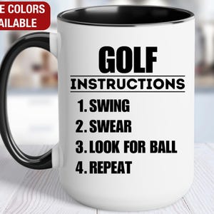 May include: White ceramic coffee mug with a black rim and handle. The mug has a black graphic that says "GOLF INSTRUCTIONS" with a list of instructions: "1. SWING", "2. SWEAR", "3. LOOK FOR BALL", "4. REPEAT".