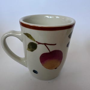 Könnte beinhalten: Ein cremefarbener Keramikbecher mit einem handgemalten Motiv eines rot-gelben Apfels mit grünen Blättern an einem Zweig. Der Becher hat einen braun bemalten Rand an der Oberseite und einen geschwungenen Henkel auf der linken Seite.