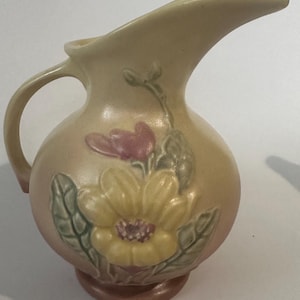 May include: A ceramic pitcher with a rounded body and a curved handle. The pitcher features a matte finish in shades of cream and pink, decorated with a raised, embossed yellow flower and green leaves on the front surface.