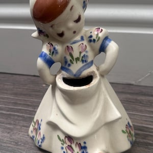 May include: A ceramic figurine of a person wearing a bonnet and a dress with a floral pattern. The figure has a brown cap, blue accents on the sleeves and bodice, and pink and blue tulip motifs on the skirt. A small opening is located at the waist.