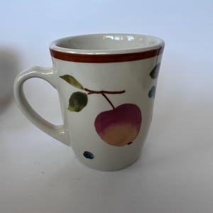 Könnte beinhalten: Ein cremefarbener Keramikbecher mit einem aufgemalten Motiv eines rot-gelben Apfels mit grünen Blättern an einem Zweig. Der Becher hat einen braunen horizontalen Streifen am oberen Rand und kleine blaue Punkte nahe dem Boden des Gefäßes.