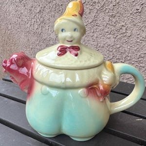 May include: A ceramic teapot shaped like a clown with a yellow hat lid, a red bow tie, and a pale green body. The spout is formed by a red pig-like figure, while a small orange animal figure is attached near the handle. The teapot features a glossy finish.