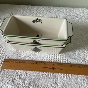 May include: Two stacked rectangular ceramic loaf pans with cream-colored glaze, green rim accents, and a Christmas tree and holly berry motif. A wooden Westcott ruler is placed in front, showing the pans are approximately 11 inches in length.