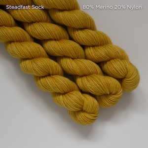 Puede incluir: Primer plano de un ovillo de hilo Steadfast Sock en color amarillo dorado. La etiqueta indica que es 80% lana merino y 20% nailon. El hilo está bien enrollado, mostrando una superficie texturizada.