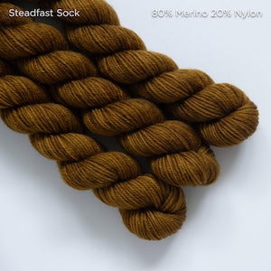 Puede incluir: Primer plano de dos madejas de hilo Steadfast Sock en un cálido color marrón dorado. La etiqueta indica que el hilo es 80% lana merino y 20% nailon. El hilo está fuertemente torcido, mostrando la textura de las fibras.