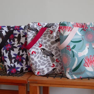 以下が含まれることがあります： カラフルな花柄と鳥柄の巾着袋が4つ。袋は木製の表面に置かれています。1つの袋は、カラフルな鳥と花が描かれた黒い背景を持っています。別の袋は、紫色の花と黒い鳥が描かれた白い背景を持っています。