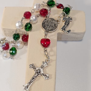 Op de afbeelding: Een wit houten kruis met een rood, wit en groen rozenkrans eroverheen gedrapeerd. De rozenkrans heeft een zilveren St. Christopher-medaille en een zilveren hartbedel. Het kruis ligt op een houten oppervlak.