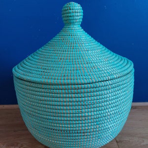 May include: A woven storage basket with a lid, featuring a turquoise blue and natural colour design. The basket is round and has a domed lid with a knob on top.