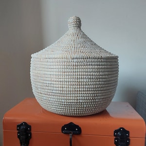 May include: A white woven basket with a lid, sitting on top of an orange metal storage trunk with black latches.