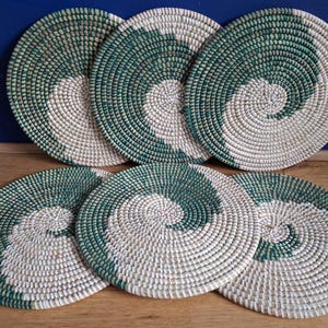 May include: Six round woven placemats. Each placemat features a spiral design with alternating sections of natural and green fibres. The placemats are arranged on a wooden surface with a blue wall in the background.