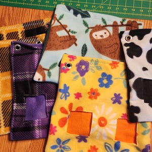 Op de afbeelding: Een verzameling kleurrijke fleece huisdieraccessoires. Ontwerpen omvatten ruitjes, luiaards, koeienprint en bloempatronen. Elk item heeft een versterkte grommet en een vierkante opening. De items zijn gerangschikt op een houten oppervlak.