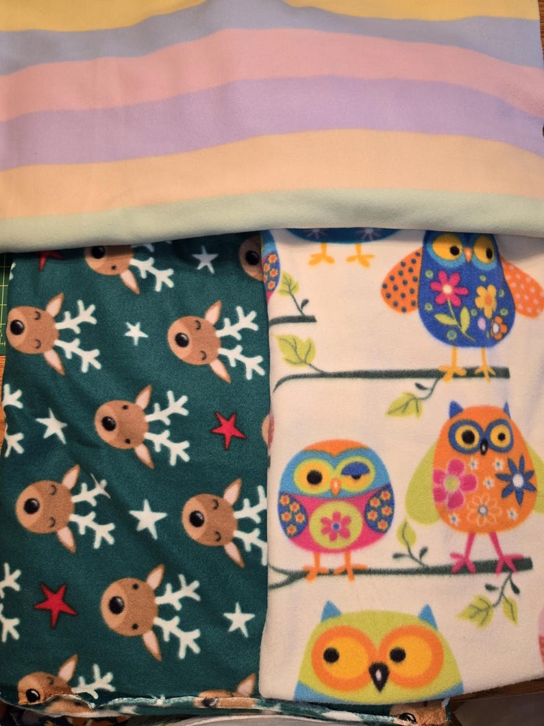 K&ouml;nnte beinhalten: Ein Stapel Fleece-Decken mit verschiedenen Mustern. Eine Decke hat pastellfarbene horizontale Streifen. Eine andere zeigt Rentiere und Schneeflocken auf teal-farbenem Hintergrund. Die dritte Decke zeigt bunte Eulen auf &Auml;sten vor wei&szlig;em Hintergrund.