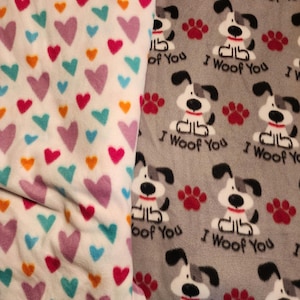 K&ouml;nnte beinhalten: Zwei Fleecedecken. Eine ist wei&szlig; mit bunten Herzen in Rosa, T&uuml;rkis, Orange und Lila. Die andere ist grau mit sich wiederholenden Bildern eines Zeichentrickhundes, roten Pfotenabdr&uuml;cken und dem Text "I Woof You."