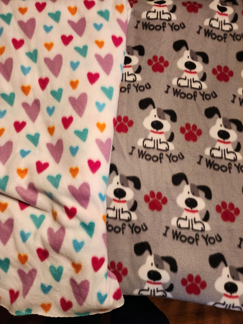 K&ouml;nnte beinhalten: Zwei Fleecedecken. Eine ist wei&szlig; mit bunten Herzen in Rosa, T&uuml;rkis, Orange und Lila. Die andere ist grau mit sich wiederholenden Bildern eines Zeichentrickhundes, roten Pfotenabdr&uuml;cken und dem Text "I Woof You."