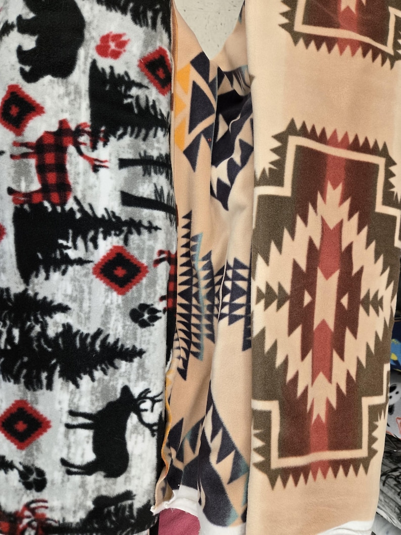 K&ouml;nnte beinhalten: Nahaufnahme von drei gerollten Fleece-Stoffen mit verschiedenen Mustern. Ein Stoff hat einen grauen Hintergrund mit schwarzen B&auml;umen, B&auml;ren, Hirschen und roten Karomustern. Ein anderer hat einen beigefarbenen Hintergrund mit geometrischen Formen in Marineblau, Gelb und Wei&szlig;. Der dritte hat einen beigefarbenen Hintergrund mit einem braunen und roten geometrischen Design.
