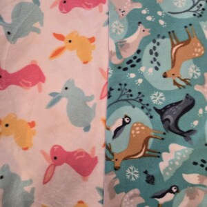 K&ouml;nnte beinhalten: Zwei Fleece-Stoffbahnen. Eine Bahn zeigt einen wei&szlig;en Hintergrund mit bunten Cartoon-Hasen in Rosa, Gelb und Blau. Die andere Bahn hat einen t&uuml;rkisfarbenen Hintergrund mit Waldtieren, darunter Hirsche, Pinguine und F&uuml;chse.
