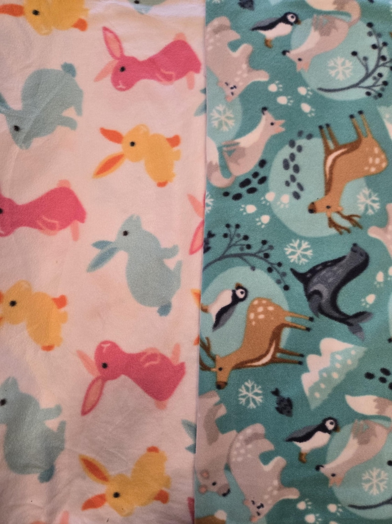 K&ouml;nnte beinhalten: Zwei Fleece-Stoffbahnen. Eine Bahn zeigt einen wei&szlig;en Hintergrund mit bunten Cartoon-Hasen in Rosa, Gelb und Blau. Die andere Bahn hat einen t&uuml;rkisfarbenen Hintergrund mit Waldtieren, darunter Hirsche, Pinguine und F&uuml;chse.