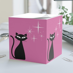 May include: A pink cube-shaped sticky note holder featuring a black cat design with starburst accents. The cat illustrations are stylized and have large, expressive eyes. The holder sits on a white surface with paperclips.