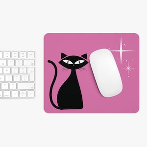 May include: A pink mousepad with a black cat silhouette and a white mouse. The cat has a stylized design with pointed ears and a long tail. A white starburst design is on the right side of the mousepad. A white keyboard is on the left.