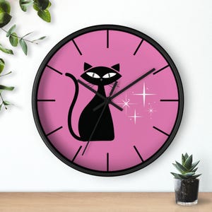 May include: A black-framed wall clock with a pink face featuring a black cat silhouette and starburst accents. The clock has black hands and hour markers. The cat has large eyes and a curved tail.