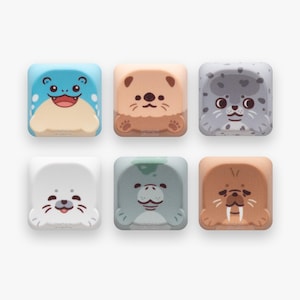 May include: A set of six square mechanical keyboard keycaps featuring illustrations of various marine animals including a blue creature, a tan otter, a spotted gray seal, a white seal, a green manatee, and a brown walrus with white tusks.