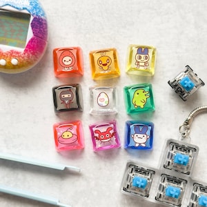 May include: A collection of colorful, square-shaped keycaps featuring various cartoon characters. The keycaps are in shades of orange, yellow, green, pink, and blue. A Tamagotchi device is visible in the upper left corner, and a keyboard switch is in the upper right.
