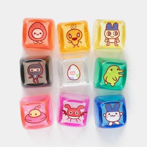 May include: Nine colourful, square-shaped, clear resin charms with cartoon character designs. The charms feature various characters in orange, yellow, green, pink, and blue. The designs include an egg, a ninja, an alien, and other cute creatures. The charms are arranged in a 3x3 grid.