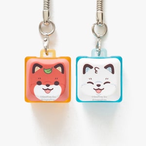 May include: Two square keyrings featuring cartoon fox faces. One is orange and red with a green leaf, the other is white and blue. Each has a silver chain and the text "©Geoffrey Siu @itsgeoffrey".