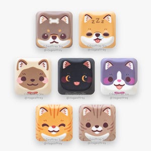 May include: Seven square keycaps with cute cartoon animal designs. The keycaps feature dogs and cats with expressive faces and bright colours. Each keycap has the artist's name and social media handle printed on it.